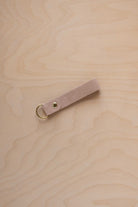 A warm beige suede leather keychain with a gold metal ring and rivet, displayed diagonally on a light wood grain background.