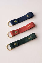 Three personalized suede keychains in navy blue, rust red, and dark green, each featuring a gold-tone metal ring and snap button with custom gold initials or lettering, arranged diagonally on a light background.