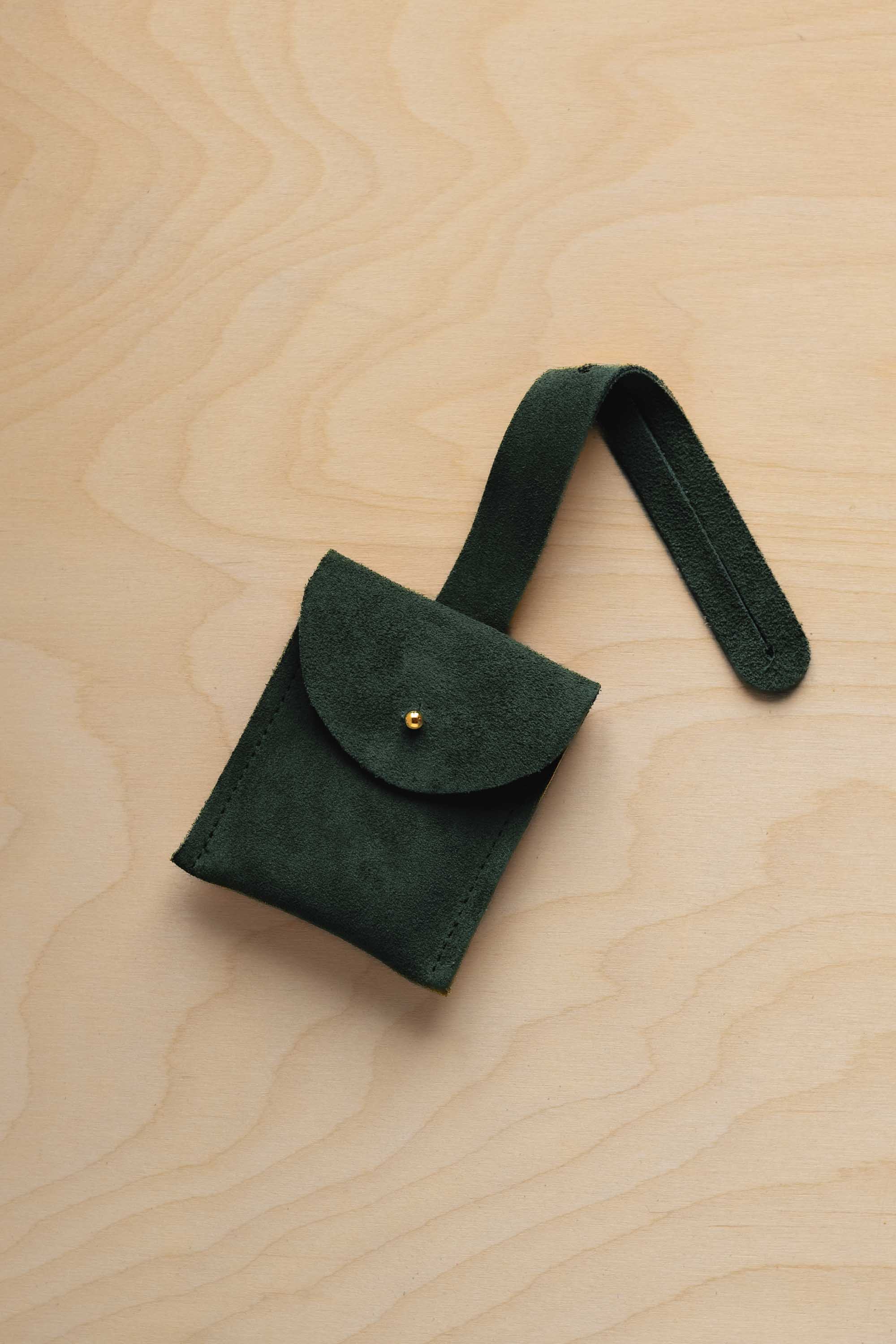 A small dep forest green suede pouch with a flap closure and a matching strap, resting on a light wood grain surface.