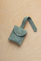 A small eucalyptus sage green suede pouch with a flap closure and a matching strap, resting on a light wood grain surface.
