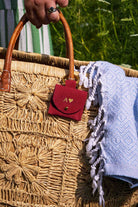 A close-up of a woven straw tote bag with a hand holding the top handle. A small red suede pouch with a gold heart logo and monogram hangs from the bag, and a light blue patterned fringed throw blanket is draped over the side.
