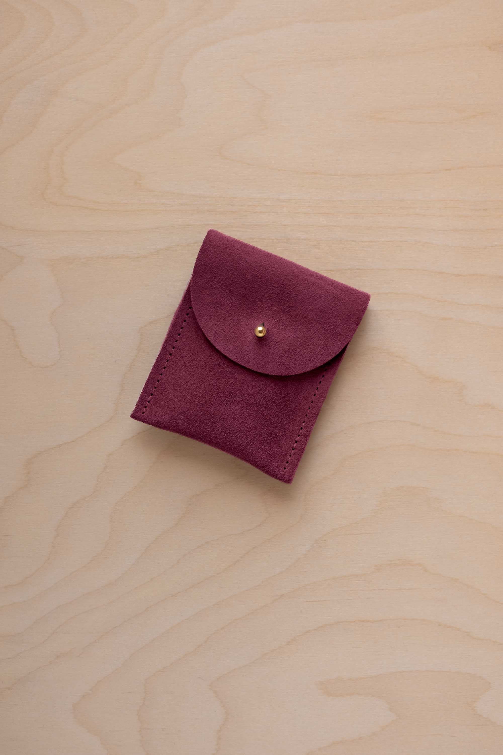 A small burgundy coloured suede pouch with a gold coloured fastening and flap closure, placed on a light wood grain surface.