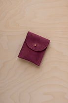 A small burgundy coloured suede pouch with a gold coloured fastening and flap closure, placed on a light wood grain surface.