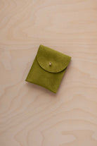 A small chartreuse green coloured suede pouch with a gold coloured fastening and flap closure, placed on a light wood grain surface.