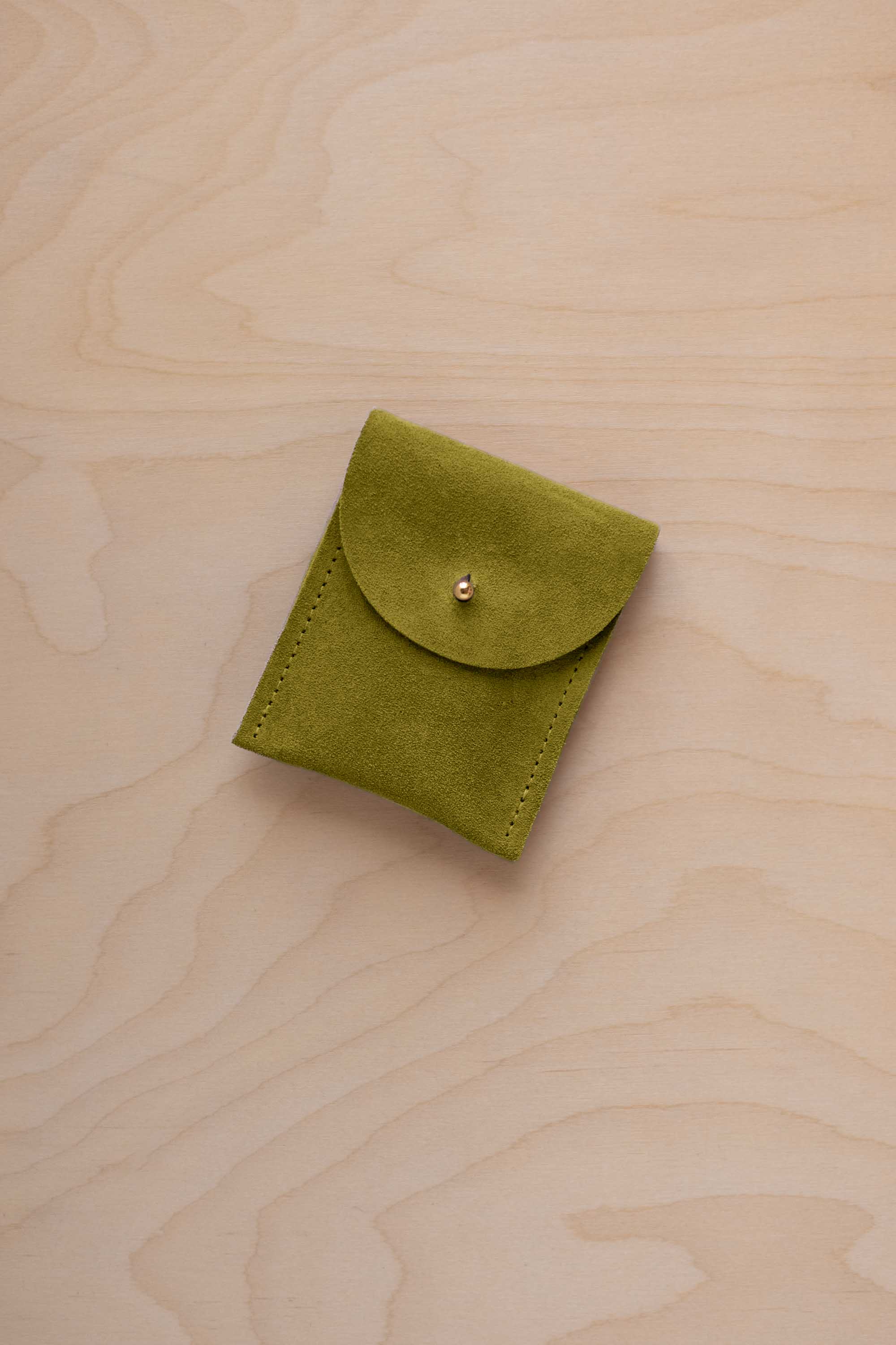 A small chartreuse green coloured suede pouch with a gold coloured fastening and flap closure, placed on a light wood grain surface.