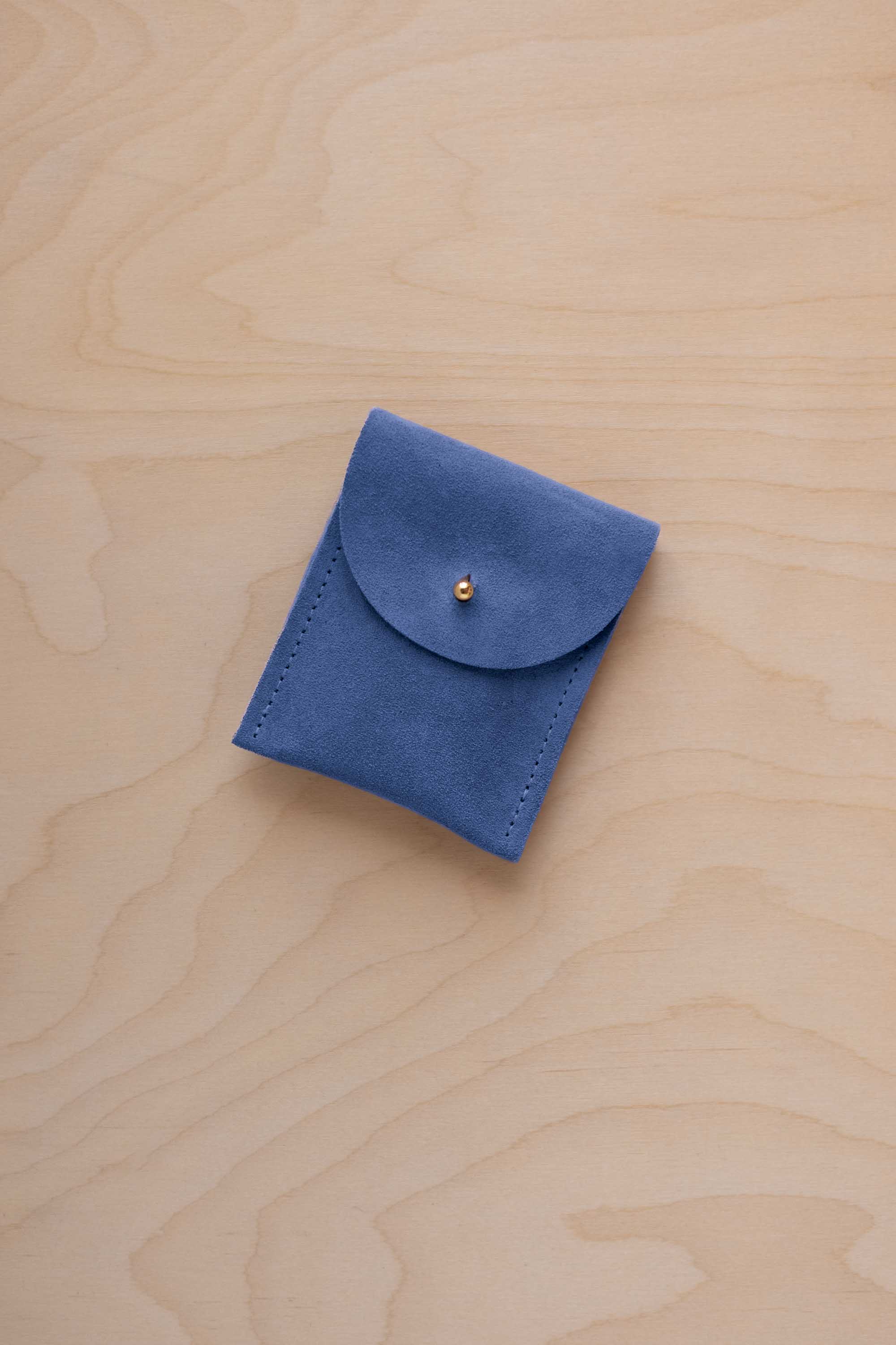 A small cobalt blue coloured suede pouch with a gold coloured fastening and flap closure, placed on a light wood grain surface.