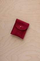 A small crimson red coloured suede pouch with a gold coloured fastening and flap closure, placed on a light wood grain surface.