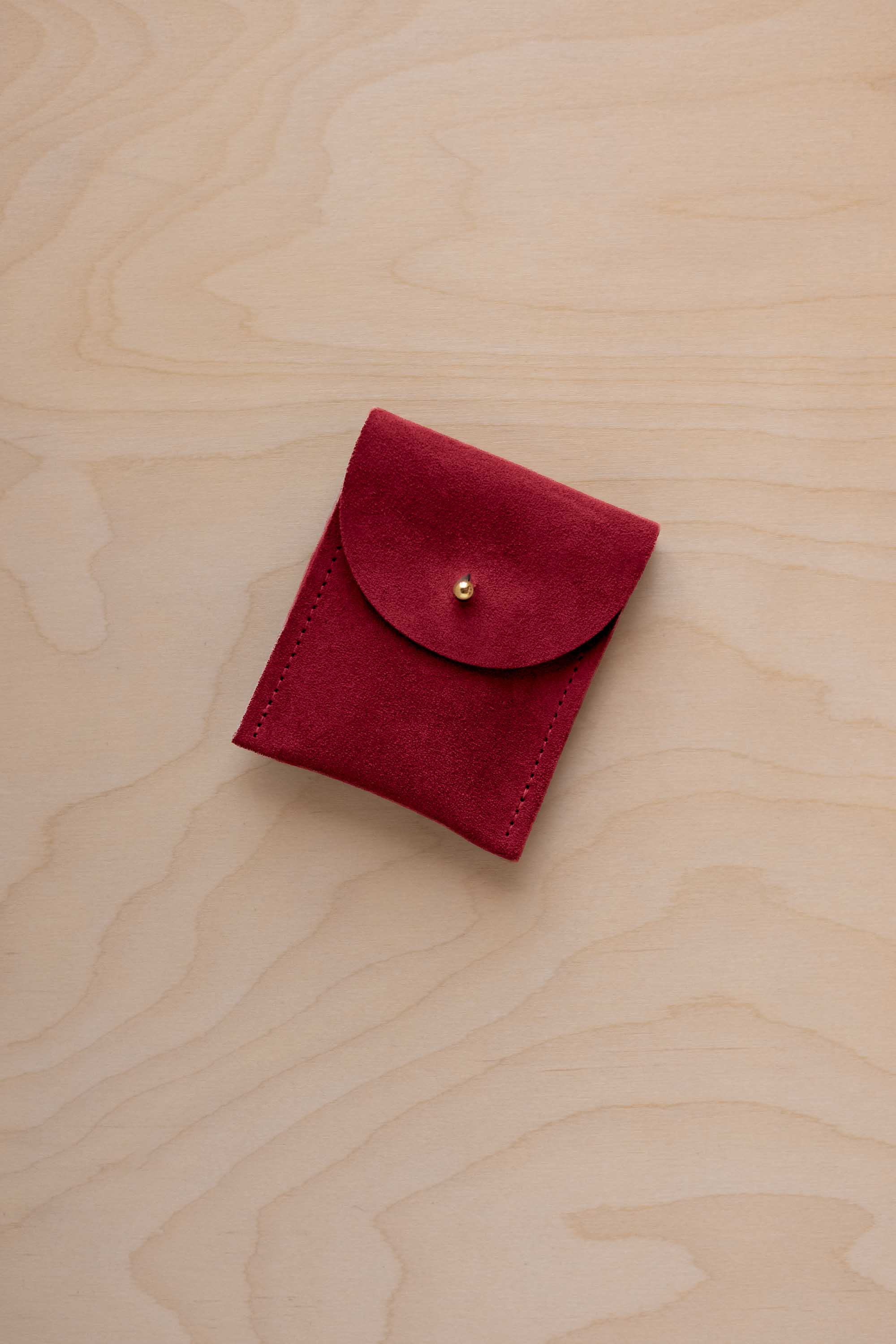 A small crimson red coloured suede pouch with a gold coloured fastening and flap closure, placed on a light wood grain surface.