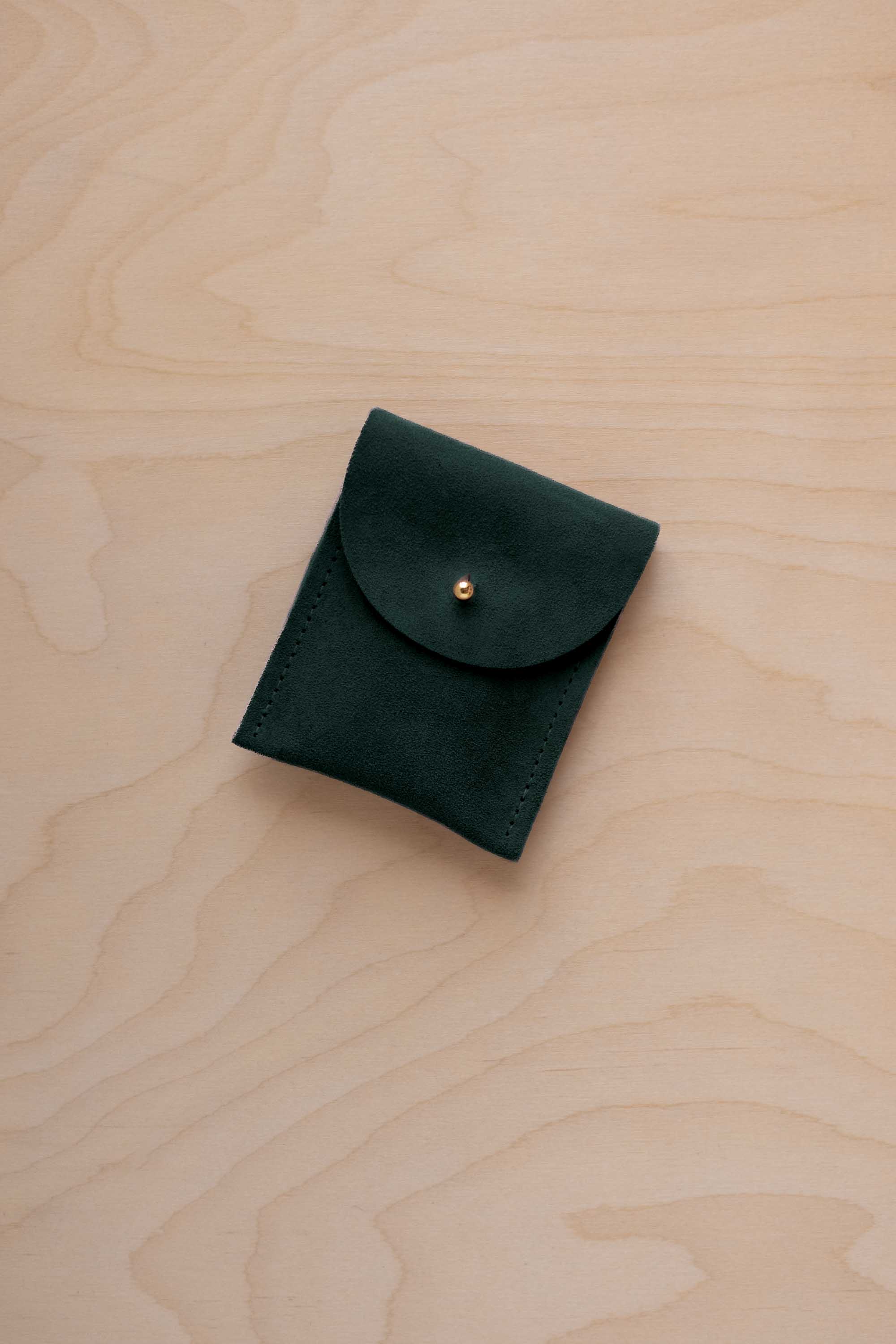 A small deep forest green coloured suede pouch with a gold coloured fastening and flap closure, placed on a light wood grain surface.