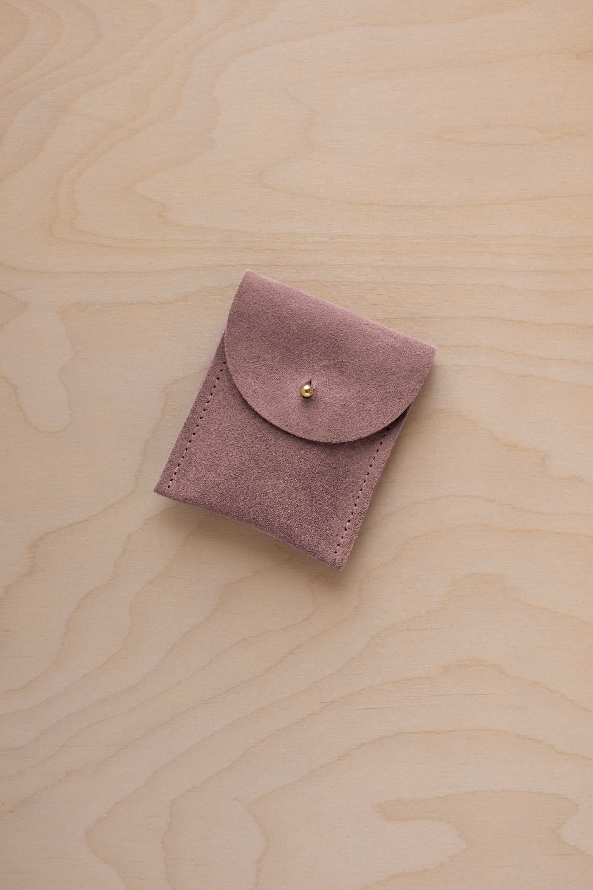 A small dusty mauve coloured suede pouch with a gold coloured fastening and flap closure, placed on a light wood grain surface.