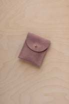 A small dusty mauve coloured suede pouch with a gold coloured fastening and flap closure, placed on a light wood grain surface.