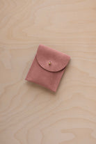 A small dusty rose coloured suede pouch with a gold coloured fastening and flap closure, placed on a light wood grain surface.