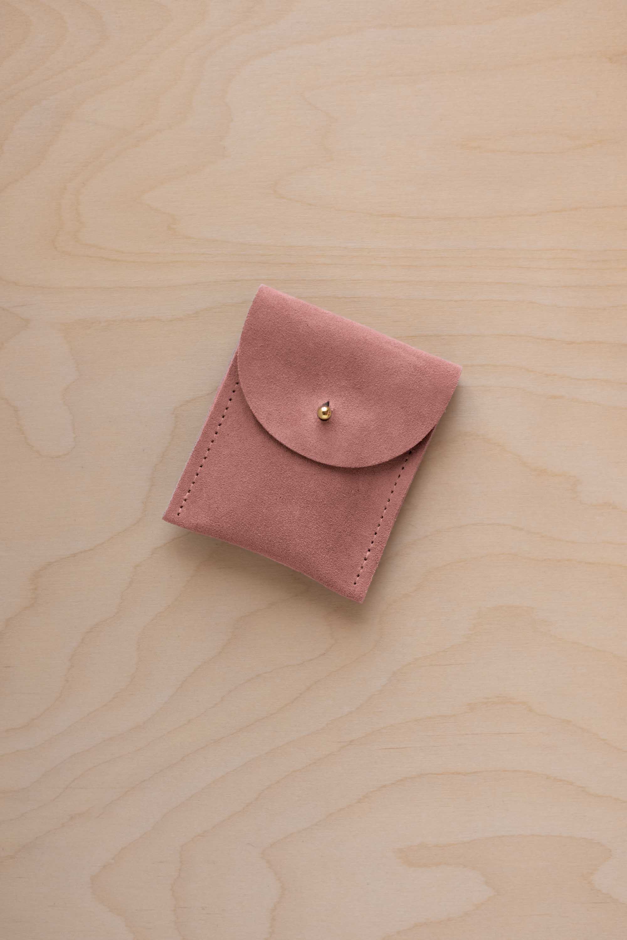 A small dusty rose coloured suede pouch with a gold coloured fastening and flap closure, placed on a light wood grain surface.