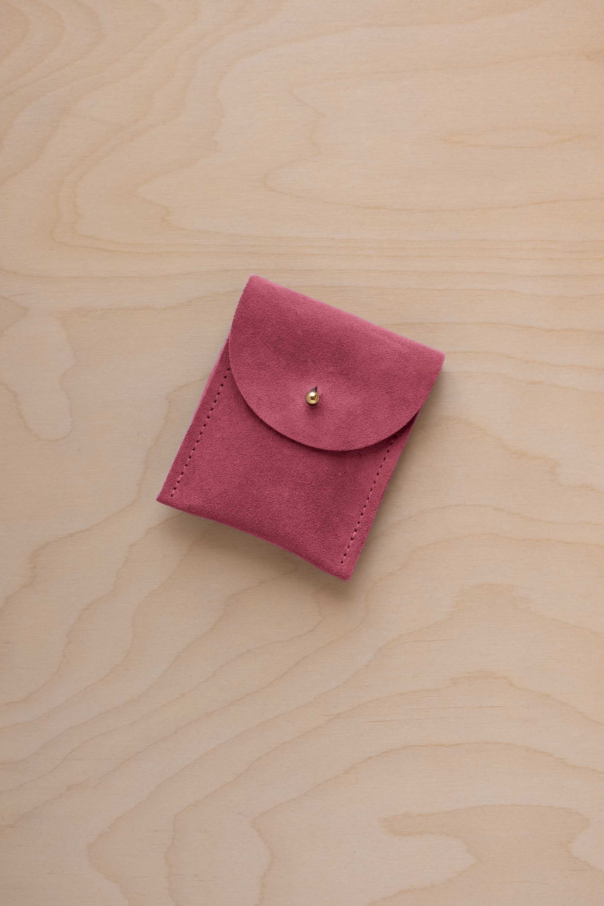 A small faded rose coloured suede pouch with a gold coloured fastening and flap closure, placed on a light wood grain surface.