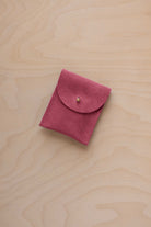A small faded rose coloured suede pouch with a gold coloured fastening and flap closure, placed on a light wood grain surface.