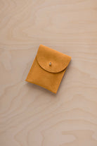 A small golden ochre coloured suede pouch with a gold coloured fastening and flap closure, placed on a light wood grain surface.
