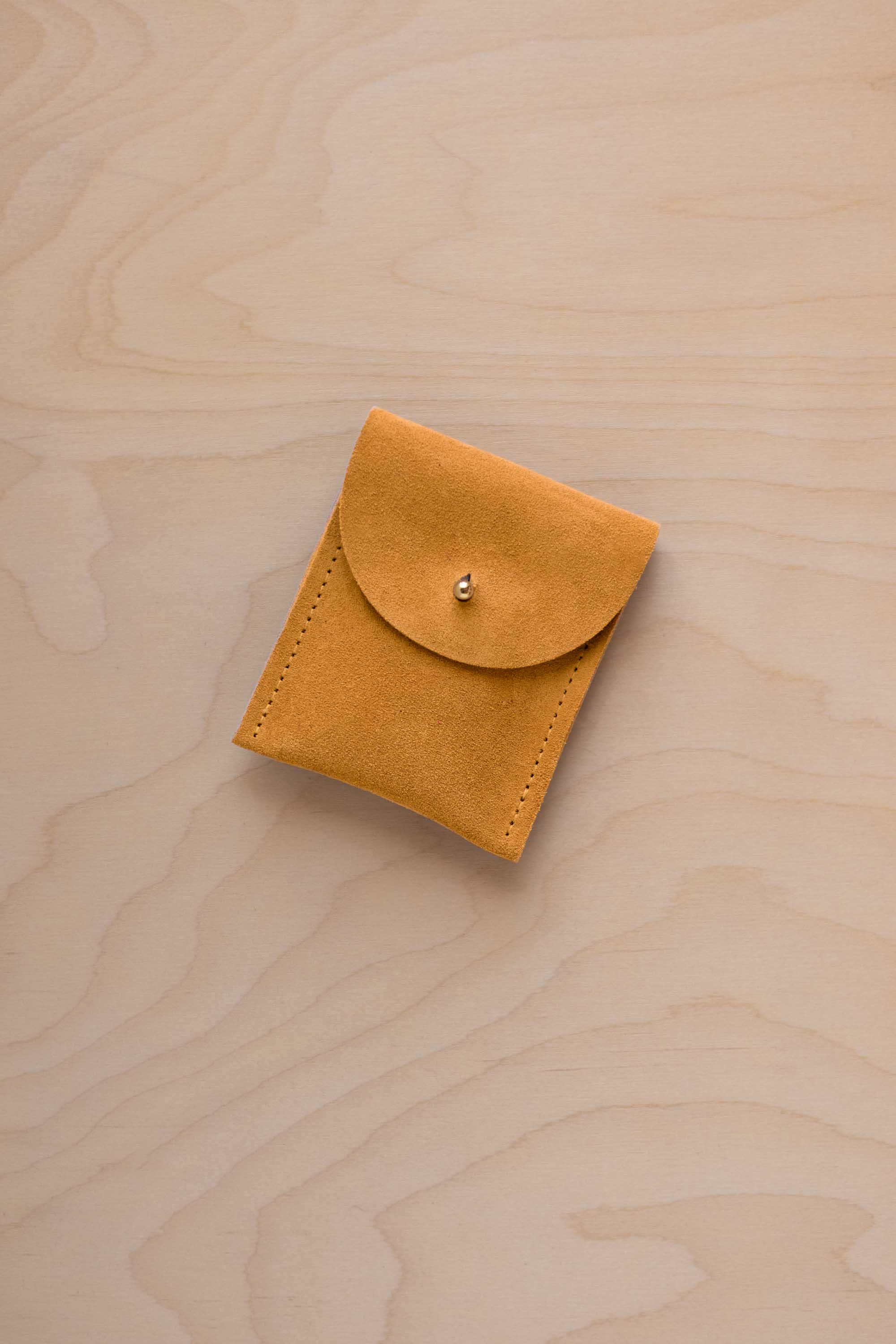 A small golden ochre coloured suede pouch with a gold coloured fastening and flap closure, placed on a light wood grain surface.