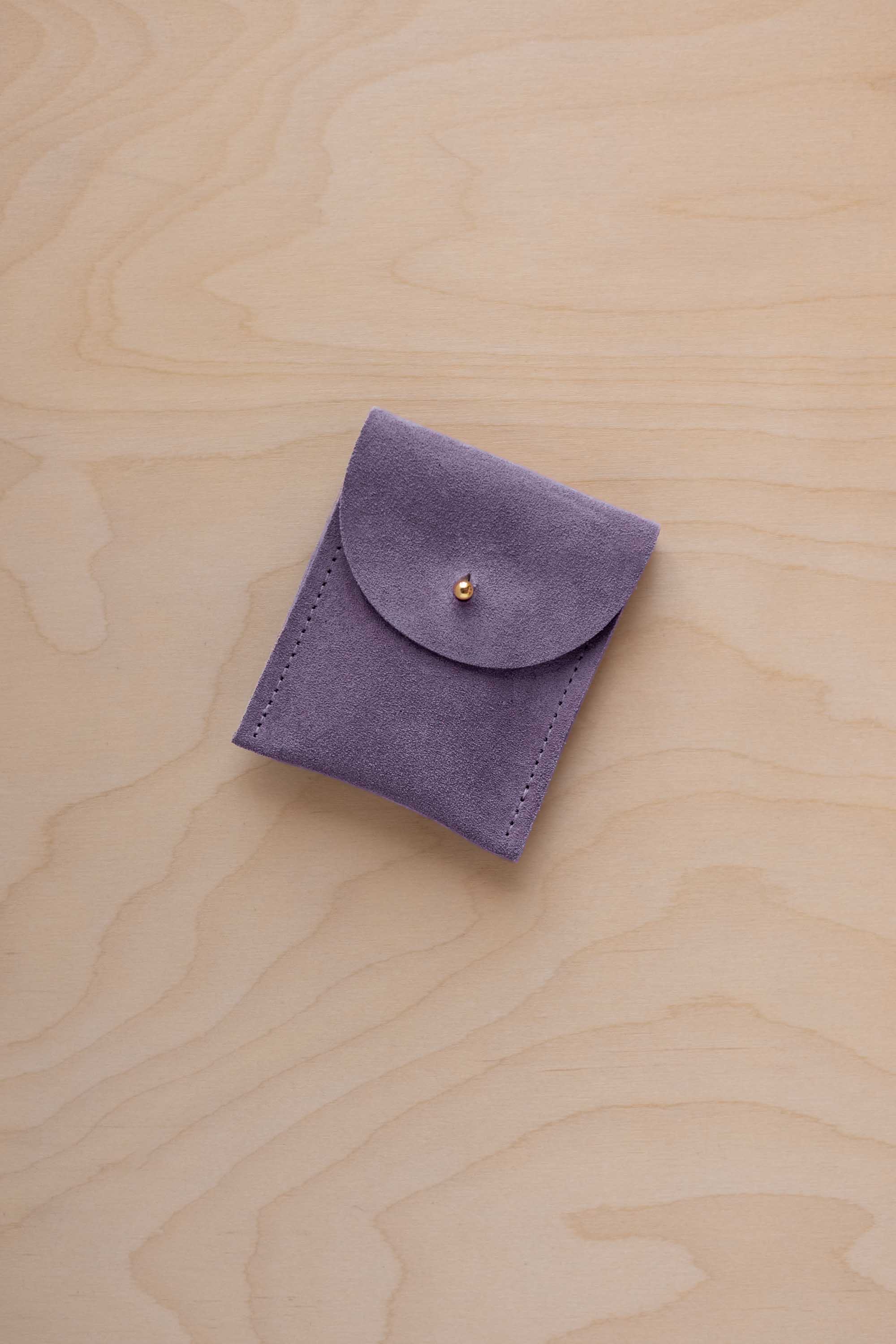 A small lavender coloured suede pouch with a gold coloured fastening and flap closure, placed on a light wood grain surface.