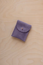 A small lavender coloured suede pouch with a gold coloured fastening and flap closure, placed on a light wood grain surface.