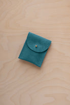 A small marine teal coloured suede pouch with a gold coloured fastening and flap closure, placed on a light wood grain surface.