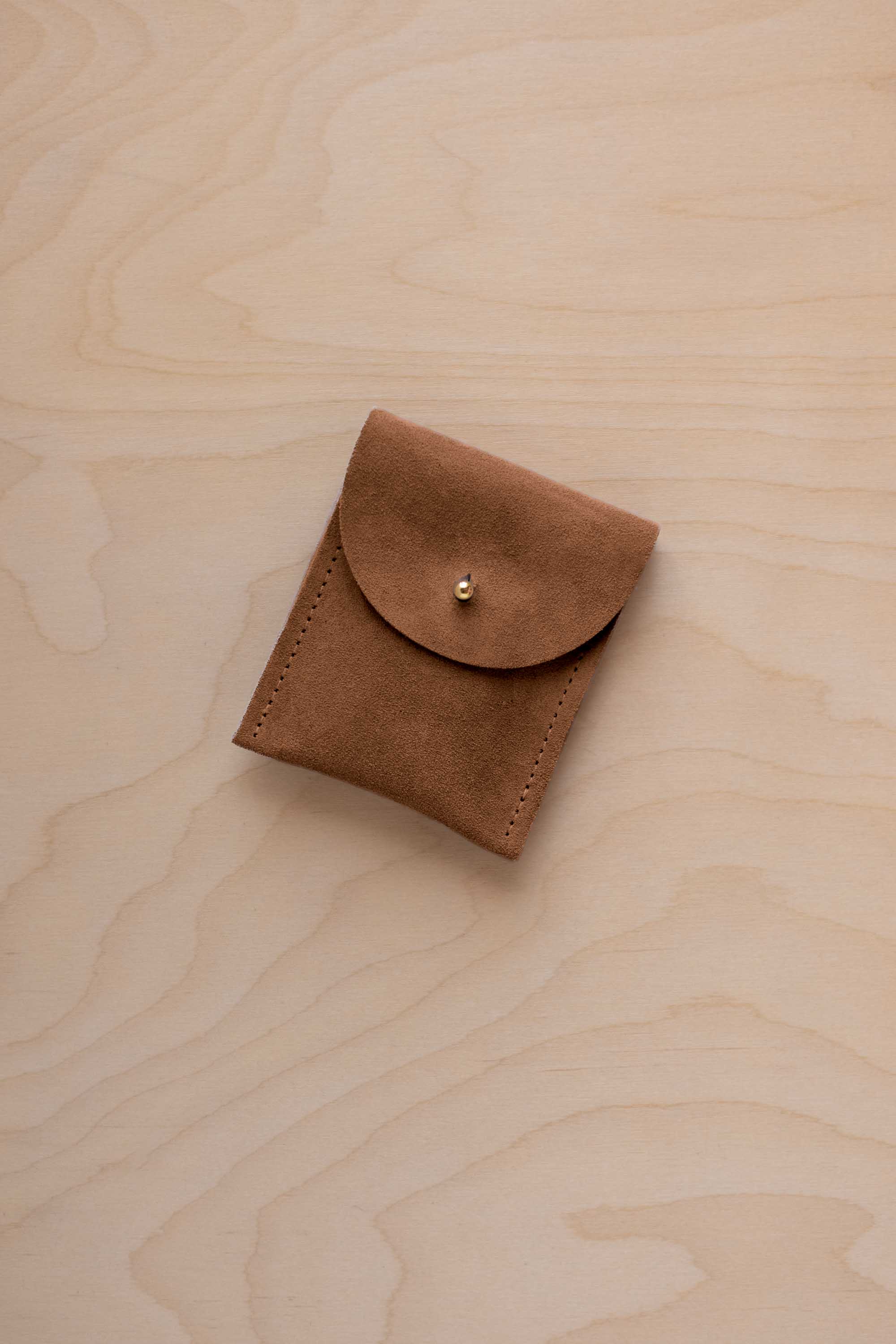 A small mocha brown coloured suede pouch with a gold coloured fastening and flap closure, placed on a light wood grain surface.