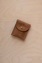 A small mocha brown coloured suede pouch with a gold coloured fastening and flap closure, placed on a light wood grain surface.