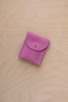 A small peony pink coloured suede pouch with a gold coloured fastening and flap closure, placed on a light wood grain surface.