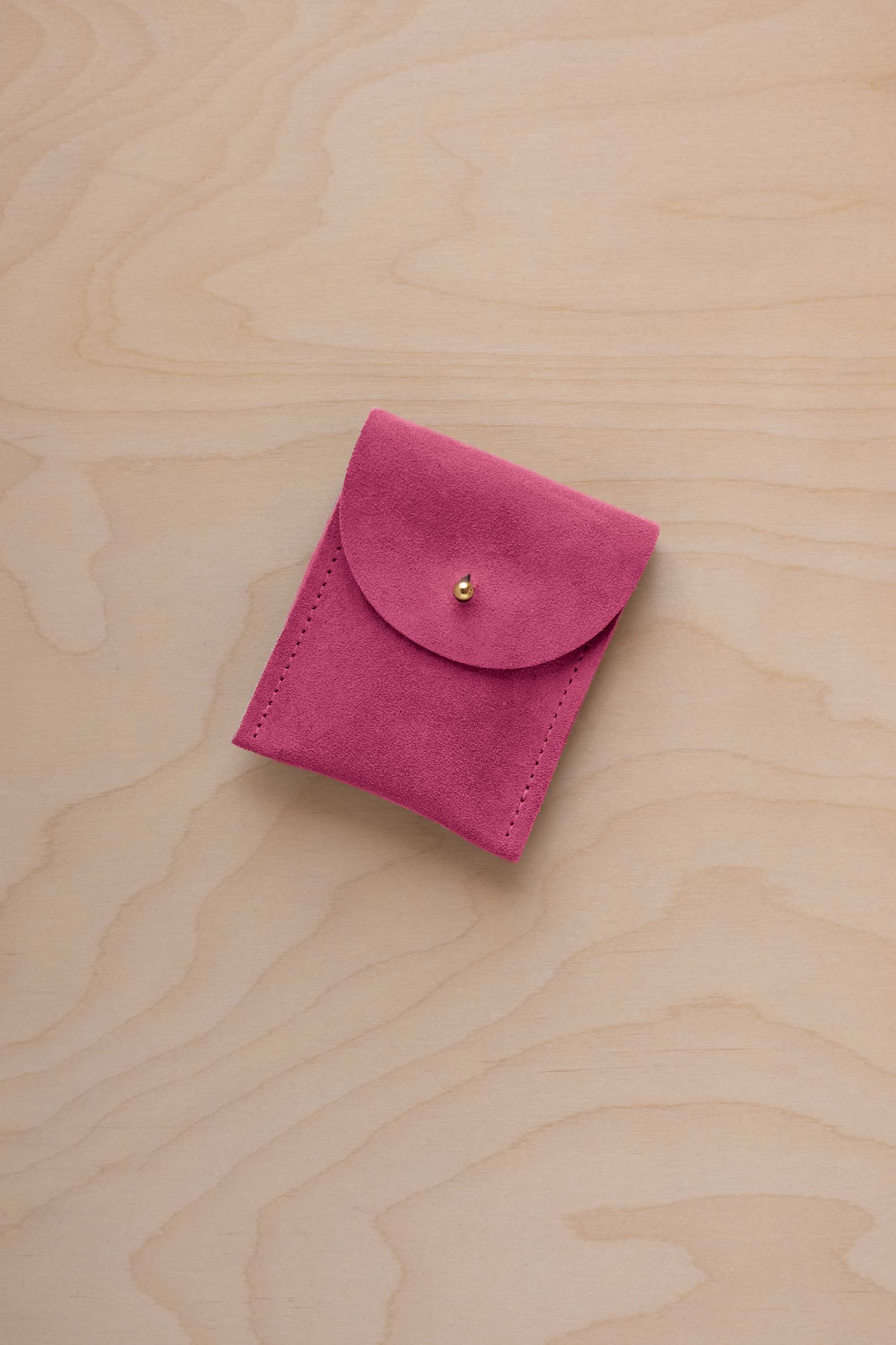 A small raspberry coloured suede pouch with a gold coloured fastening and flap closure, placed on a light wood grain surface.