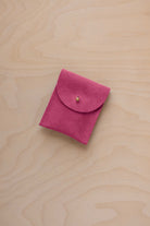 A small raspberry coloured suede pouch with a gold coloured fastening and flap closure, placed on a light wood grain surface.