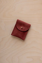 A small rust red coloured suede pouch with a gold coloured fastening and flap closure, placed on a light wood grain surface.