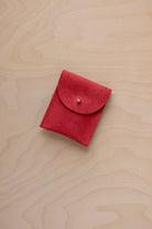 A small scarlet red coloured suede pouch with a gold coloured fastening and flap closure, placed on a light wood grain surface.