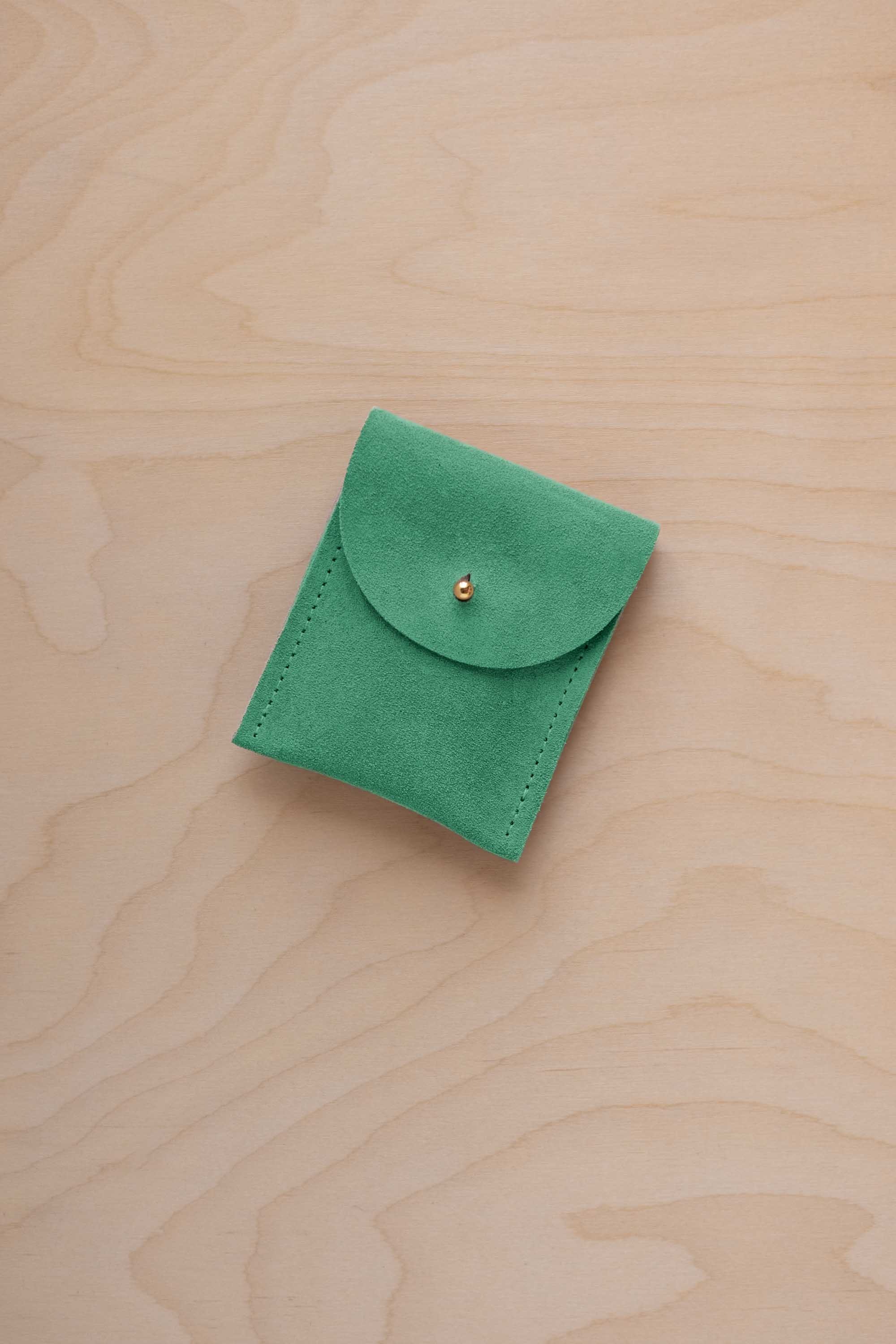 A small seafoam green coloured suede pouch with a gold coloured fastening and flap closure, placed on a light wood grain surface.
