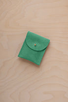 A small seafoam green coloured suede pouch with a gold coloured fastening and flap closure, placed on a light wood grain surface.