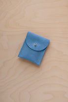 A small sky blue coloured suede pouch with a gold coloured fastening and flap closure, placed on a light wood grain surface.