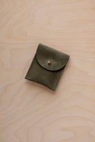 A small soft olive green coloured suede pouch with a gold coloured fastening and flap closure, placed on a light wood grain surface.
