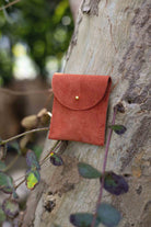 A small terracotta coloured suede pouch with a gold coloured fastening and flap closure, leaning against a natural wooden surface with an outdoor background.