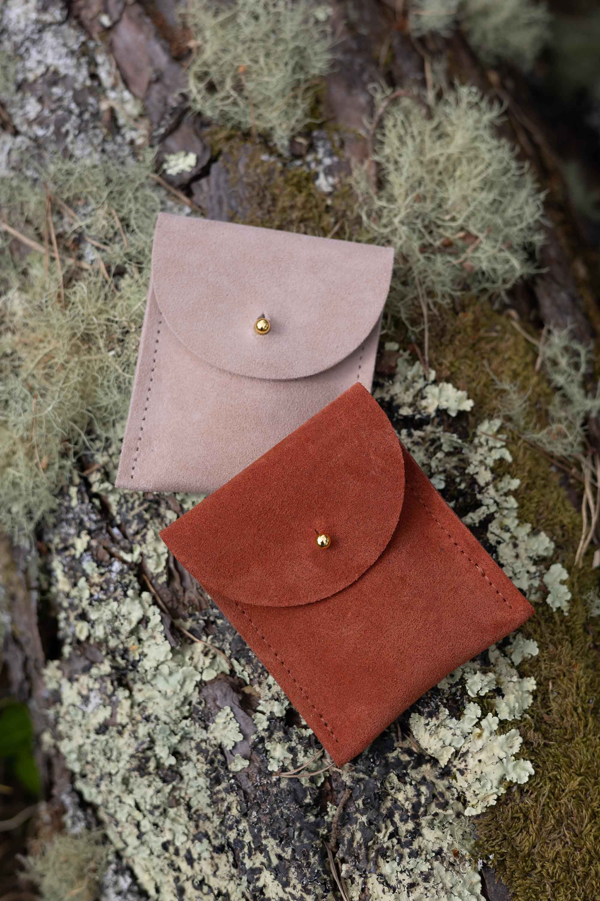 Two small coloured suede pouches with a flap closure and a gold coloured fastening, on a natural outdoor background.