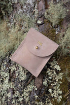 A top-down view of a warm sand coloured  suede pouch with a flap closure and a gold coloured fastening, on a natural outdoor background.