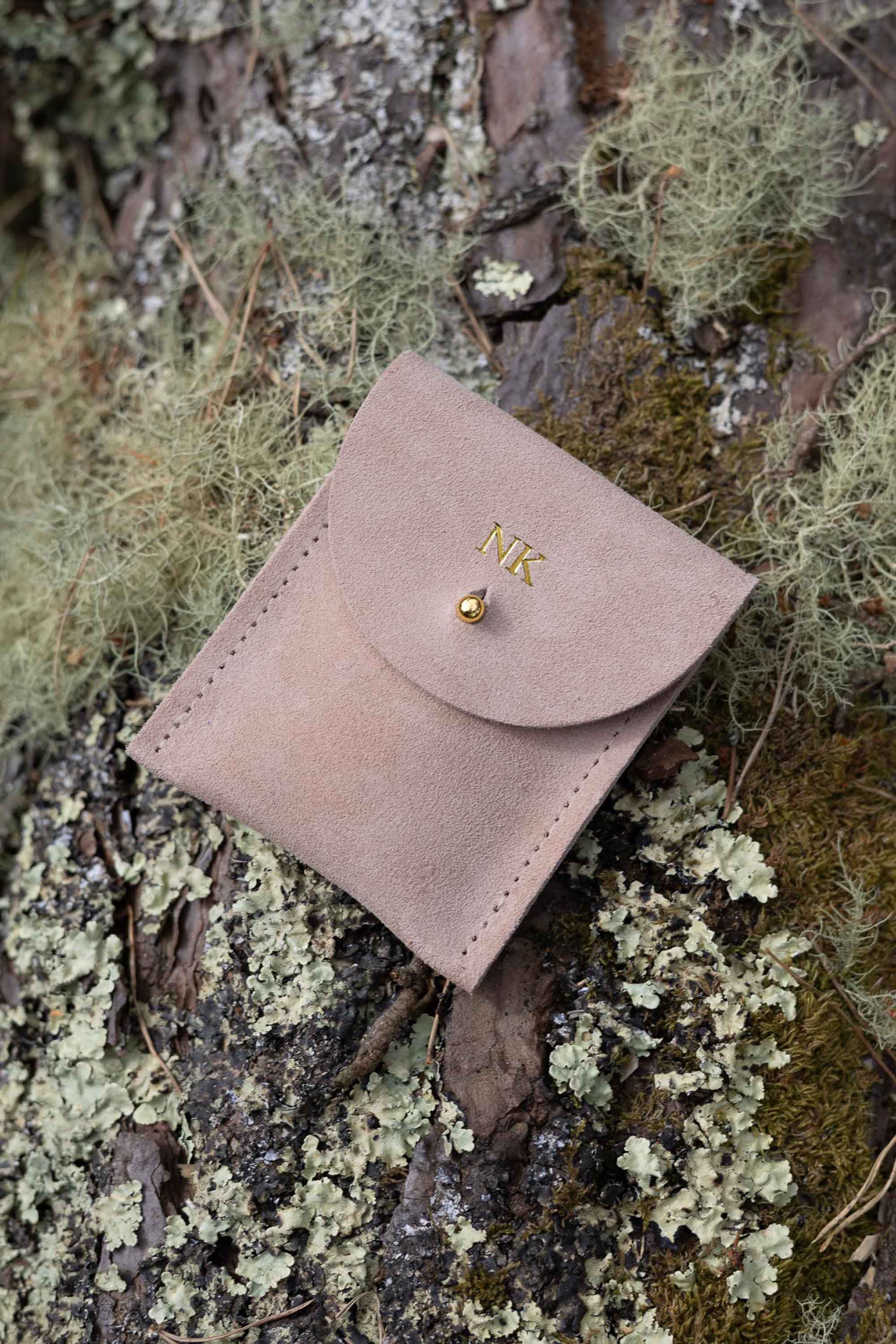 A top-down view of a warm sand coloured  suede pouch with a flap closure and a gold coloured fastening, on a natural outdoor background.