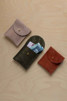 Three small suede pouches in assorted colours with a flap closure and a gold coloured fastening, arranged in a row on a light wood grain surface. The middle pouch has a bank card and a bank note nestled inside.