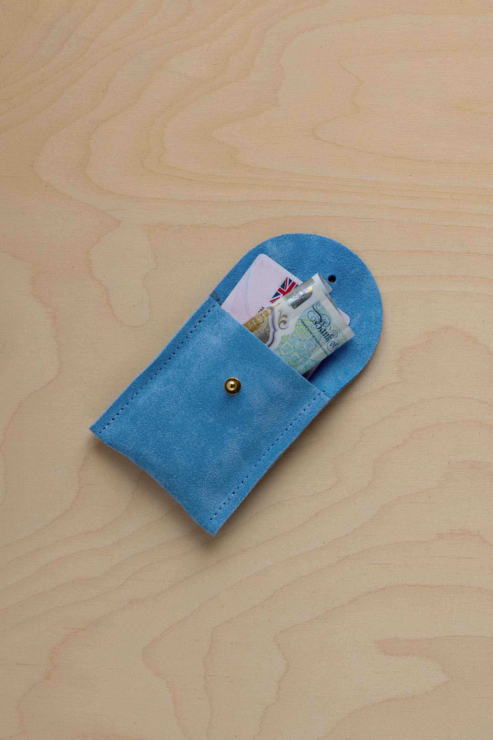 A small sky blue coloured suede pouch with a gold coloured fastening and flap closure, placed on a light wood grain surface, with a bank card and a bank note nestled inside.
