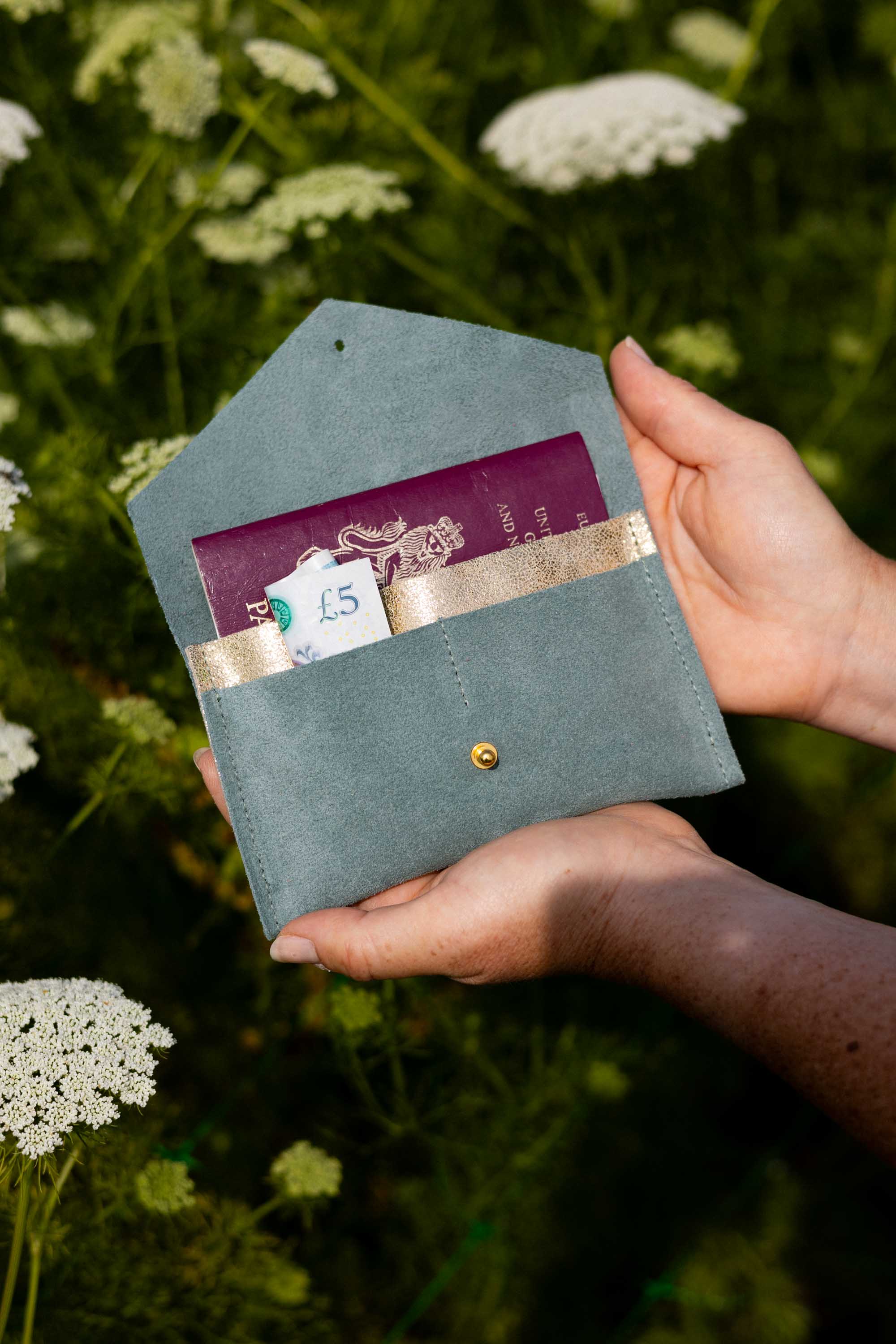 A person's hands holding an open eucalyptus or sage green envelope-style wallet with a gold-tone button, showing a purple passport and cards inside, set against a blurred background of green foliage and white wildflowers.