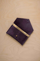 A blackcurrant dark purple suede pouch with a metallic gold contrast pocket and a small gold stud, displayed on a light wood grain background.