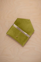 A chartreuse lime green coloured suede pouch with a metallic gold contrast pocket and a small gold stud, displayed on a light wood grain background.