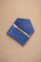 A cobalt blue suede pouch with a metallic gold contrast pocket and a small gold stud, displayed on a light wood grain background.
