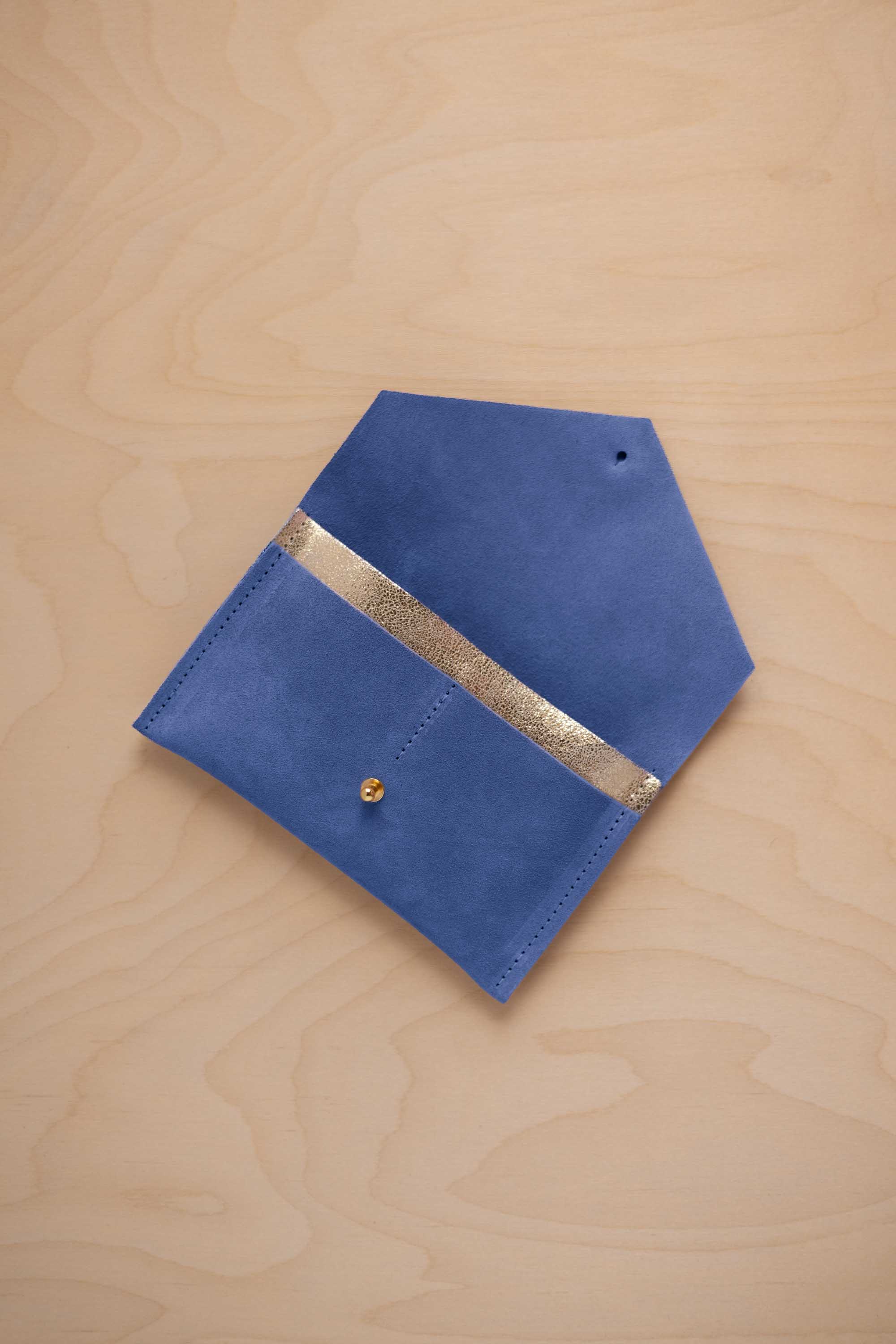 A cobalt blue suede pouch with a metallic gold contrast pocket and a small gold stud, displayed on a light wood grain background.