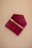 A crimson red suede pouch with a metallic gold contrast pocket and a small gold stud, displayed on a light wood grain background.