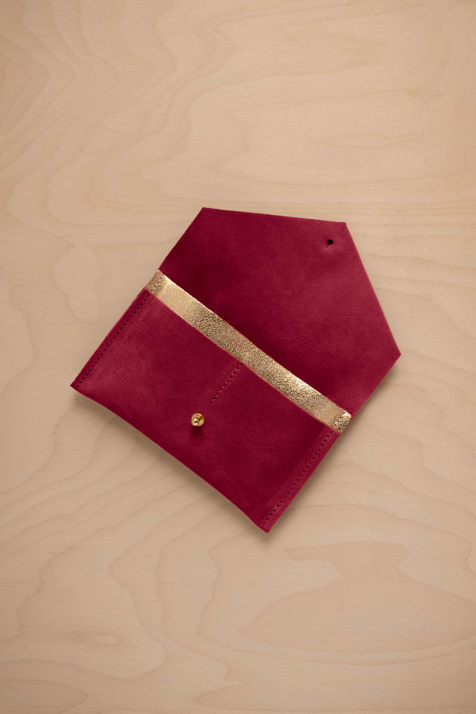 A crimson red suede pouch with a metallic gold contrast pocket and a small gold stud, displayed on a light wood grain background.