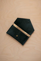 A deep forest green coloured suede pouch with a metallic gold contrast pocket and a small gold stud, displayed on a light wood grain background.