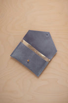 A wdusky blue suede pouch with a metallic gold contrast pocket and a small gold stud, displayed on a light wood grain background.